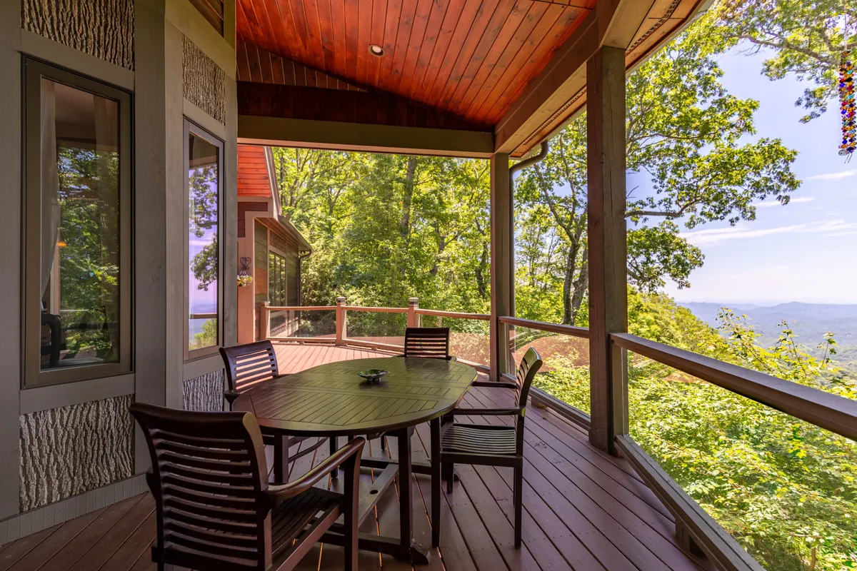 A wooden deck with an outdoor dining table set amidst surrounding trees, offering clear views of distant mountains. The table is arranged for al fresco meals, and the deck is enclosed by railings, providing a safe area for children and pets. The setting is bathed in natural light, ideal for enjoying sunrises, sunsets, and stargazing.