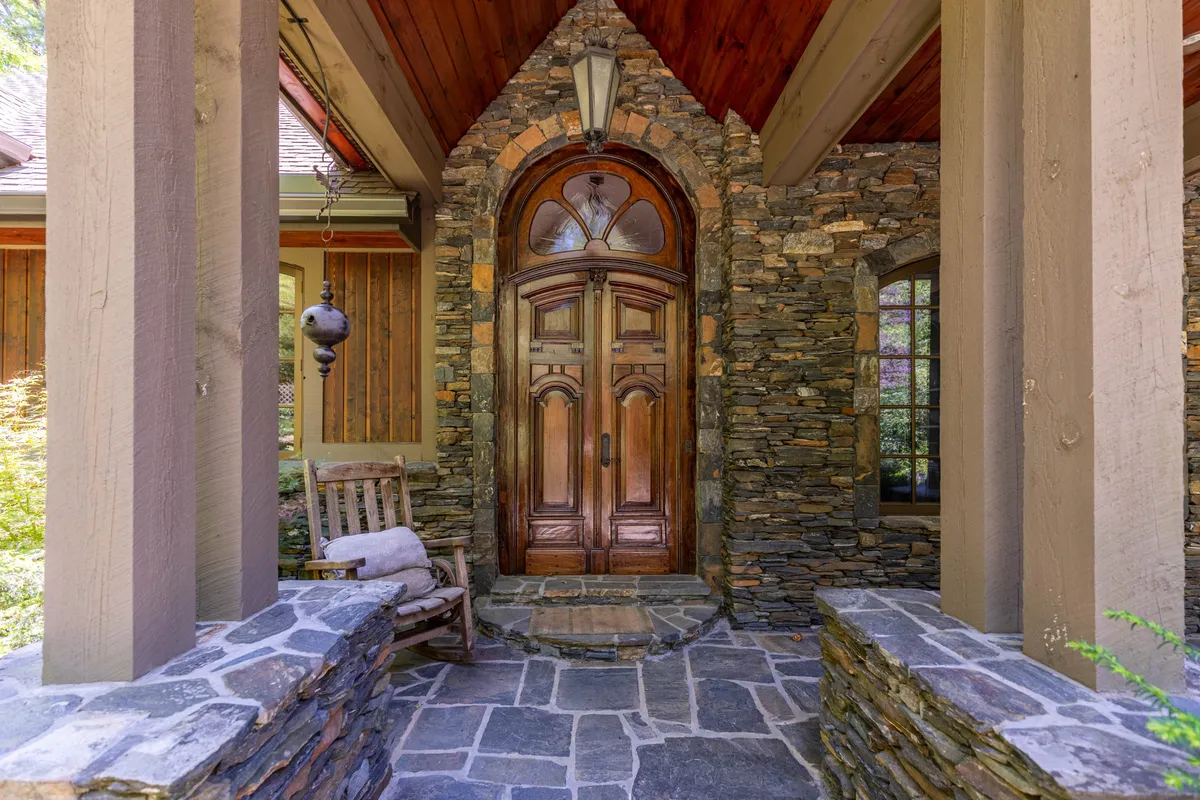 A charming exterior view of the home’s entrance, featuring an ornate wooden door framed by a striking stone archway. A wooden rocking chair sits on the porch, nestled beside a rock wall, inviting guests to relax and take in the surroundings.