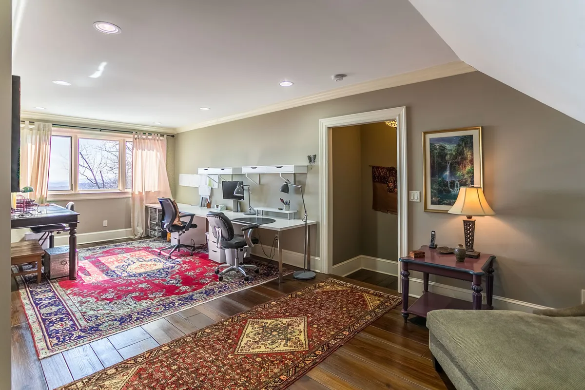 A comfortable, focused interior workspace with desks and ergonomic chairs, a large window flooding the room with natural light, and a colorful area rug. Artwork on the wall and greenery outside the window enhance the creative mood.