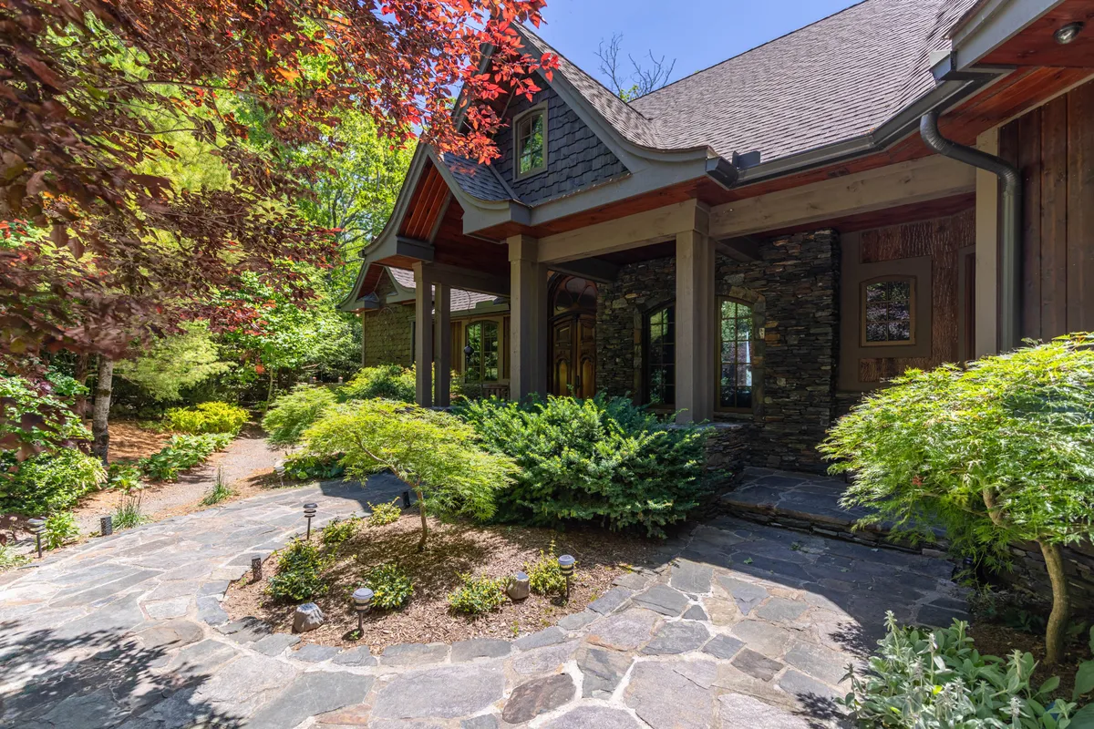 The image showcases the elegant stone facade of a custom-designed single family home, accented with warm wood details and surrounded by a beautifully landscaped garden. A charming cobblestone pathway leads up to the entrance, inviting guests to stroll through lush greenery, while a spacious porch offers panoramic sunset views over the Yadkin River Gorge and Blue Ridge Mountains.