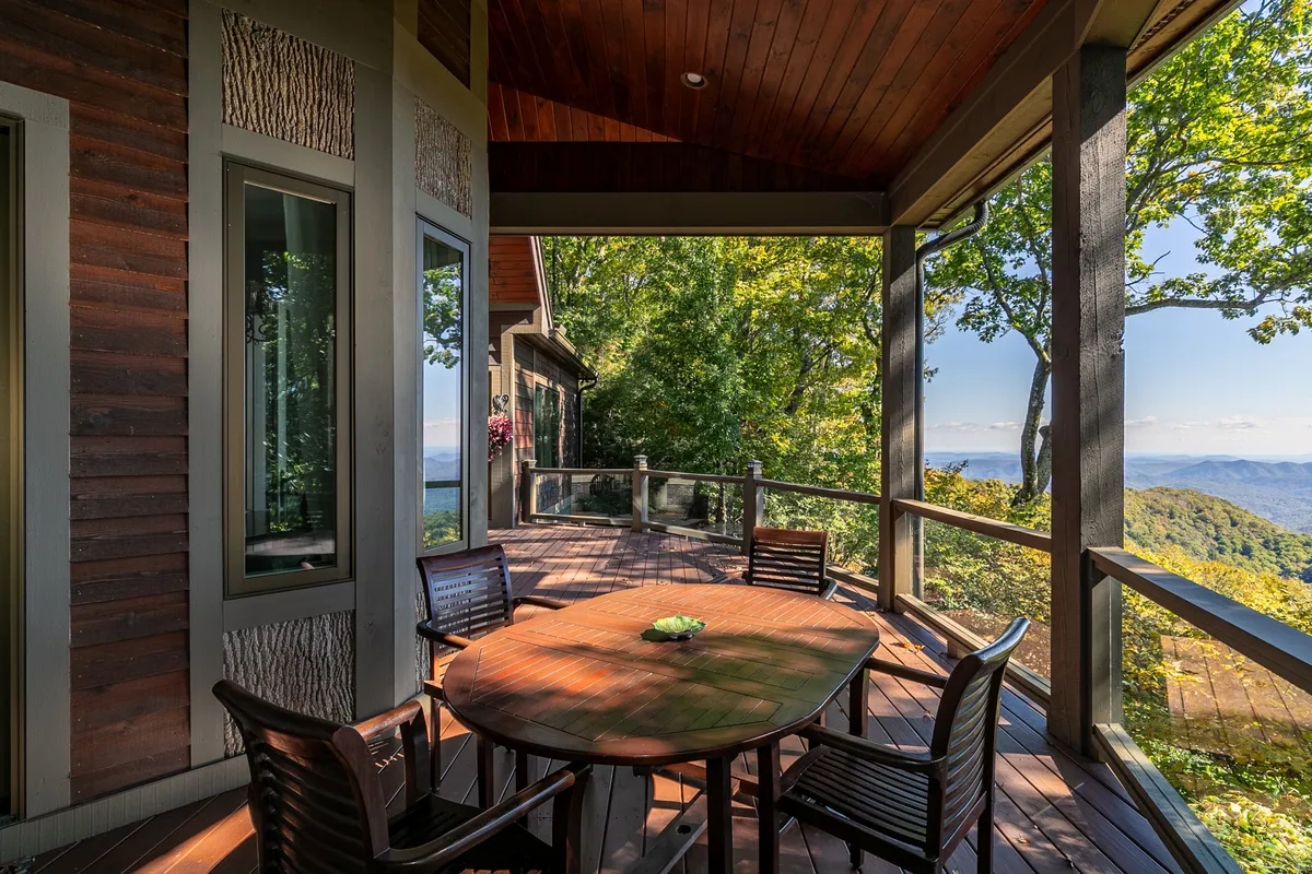 This image features a round wooden table with four chairs set on a deck, framed by green foliage and offering a sweeping view of the mountains. The setting is inviting and suggests a space ideal for outdoor dining or group gatherings, with direct access to the outdoors and scenic vistas.