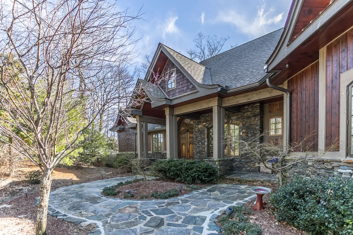 This exterior image features a welcoming stone pathway leading up to a wooden front door, framed by landscaped gardens and mature trees. The inviting porch and the surrounding greenery evoke a sense of outdoor living and relaxation.