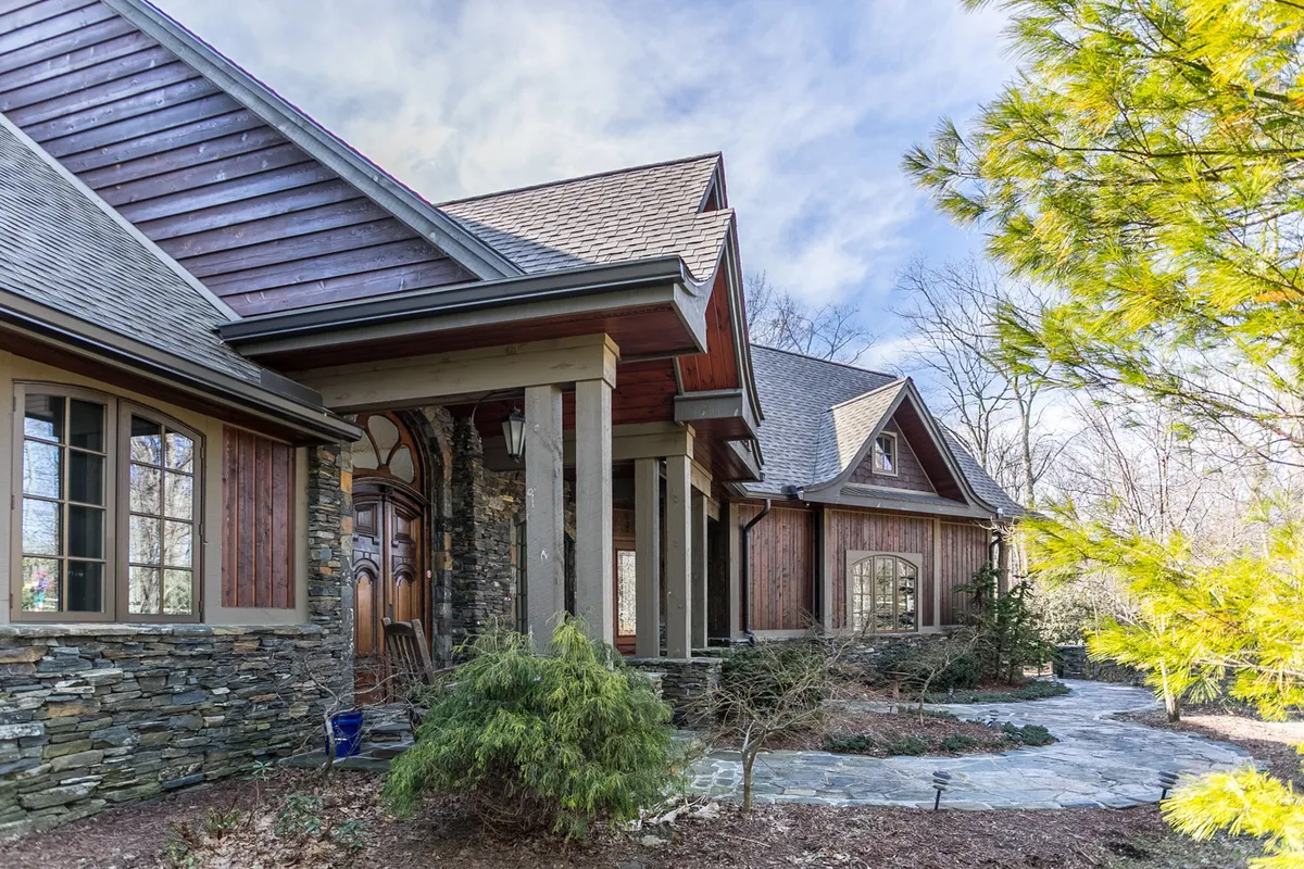 A stone facade with wooden accents, lush landscaping, and a welcoming front entrance create a warm, inviting scene. The porch and surrounding trees suggest a place for outdoor relaxation, nature appreciation, and family or pet-friendly activities.