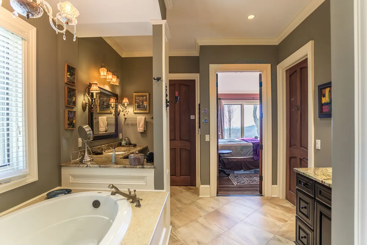 A spa-inspired ensuite bath with a freestanding bathtub, granite vanity with mirror, and a doorway connecting to the bedroom. The ambiance is serene and plush, ideal for self-care and quiet indulgence.