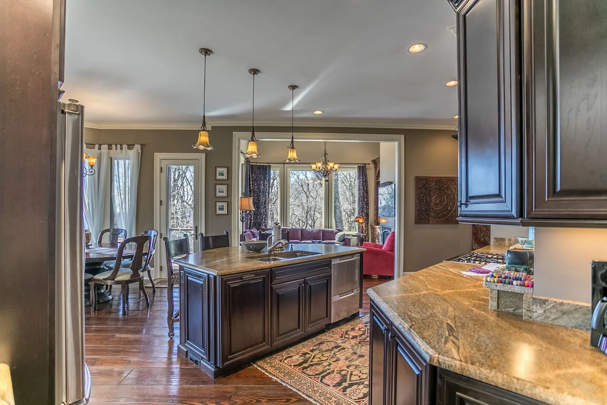 An open-concept kitchen and dining area featuring a spacious kitchen island, modern cabinetry, and large windows that bathe the space in natural light. The kitchen boasts high-end stainless-steel appliances and a welcoming dining area perfect for family meals and entertaining guests. Comfortable living room seating is visible in the background, emphasizing the flow between cooking, dining, and relaxing spaces.