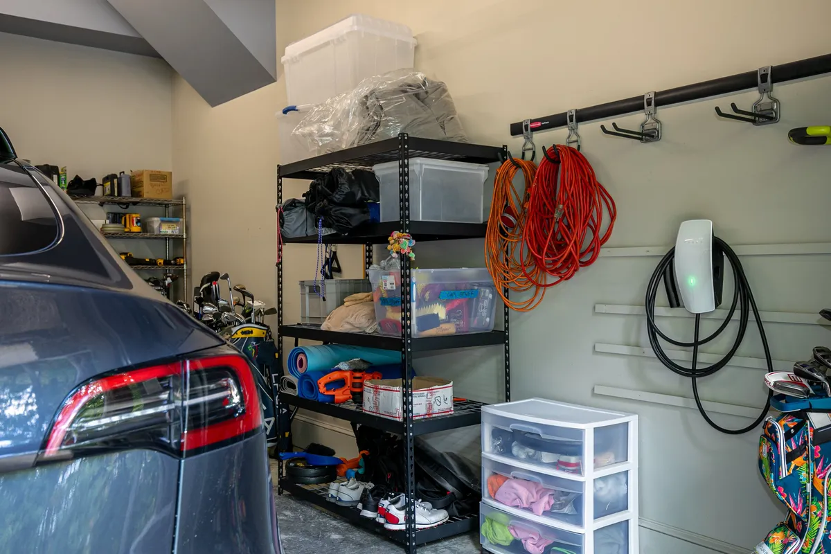 A garage space featuring an electric car parked near a wall, with visible coiled charging cords on display. The area includes organized shelving and storage bins, indicating a practical setup for vehicle charging and equipment storage.