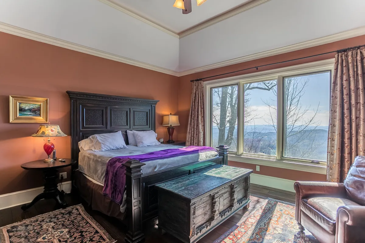 A tranquil master suite featuring a king-sized bed with a decorative headboard, large windows showcasing scenic views, a cozy armchair, and a decorative lamp on the bedside table. The space exudes calm and luxury, perfect for winding down or enjoying a morning coffee.