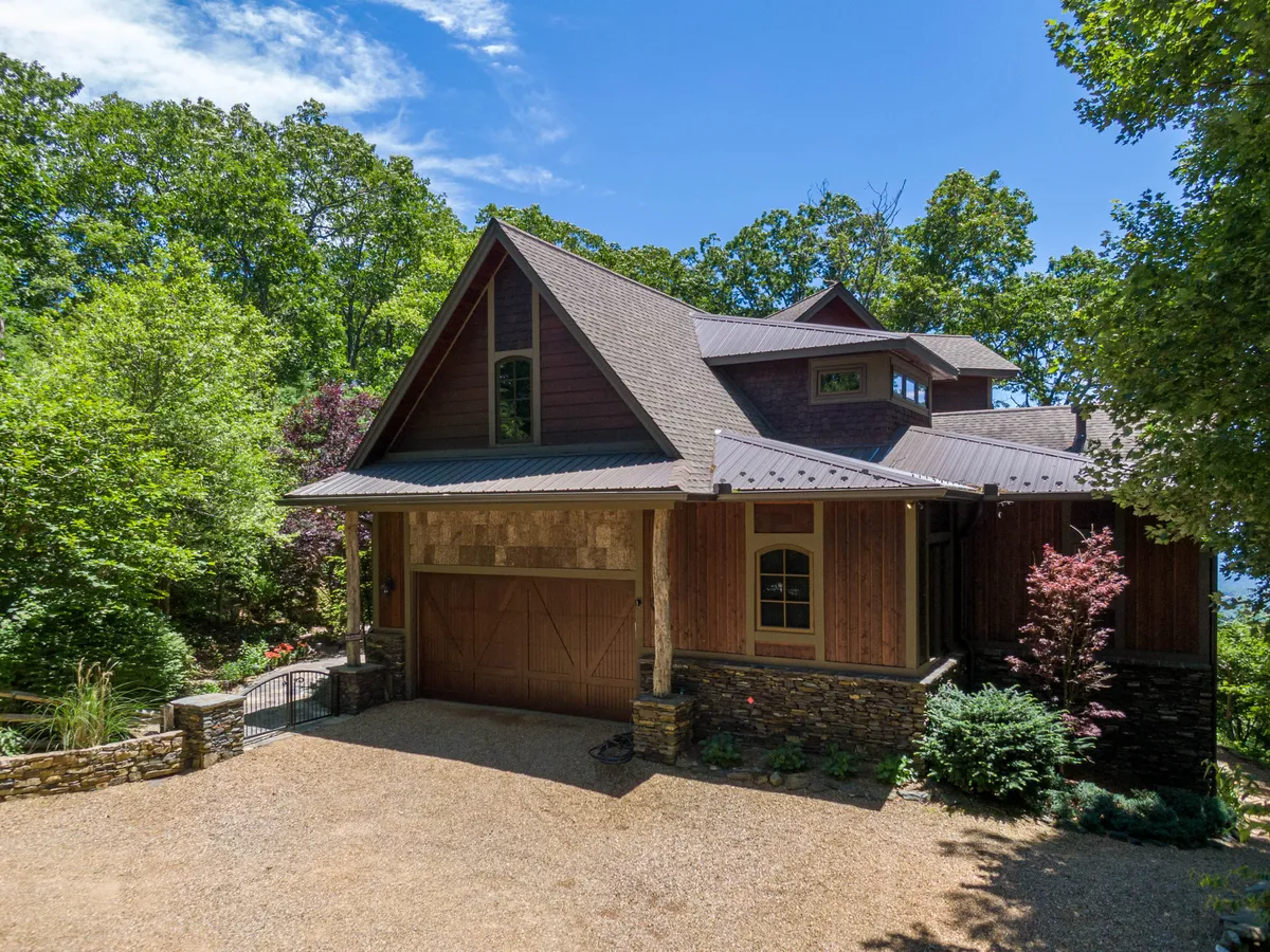 Exterior view of the house showcasing its wooden facade, a solid stone base, and a visible garage door, all surrounded by lush greenery. The inviting scene suggests ample outdoor parking and easy access for vehicles.