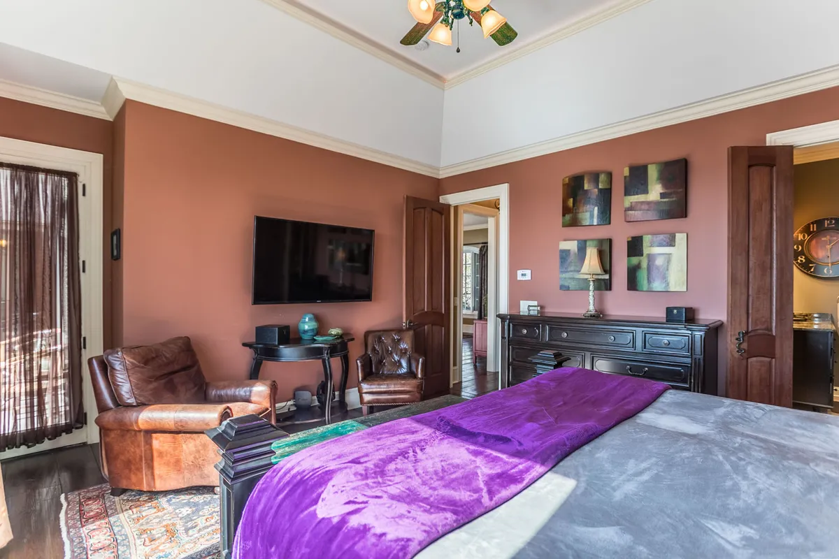 A queen bedroom with a soft purple throw on the bed, leather armchair, wall-mounted TV, and tasteful artwork. The room feels tranquil and inviting, designed for restful nights or private retreat. Natural light filters in, enhancing the peaceful mood.