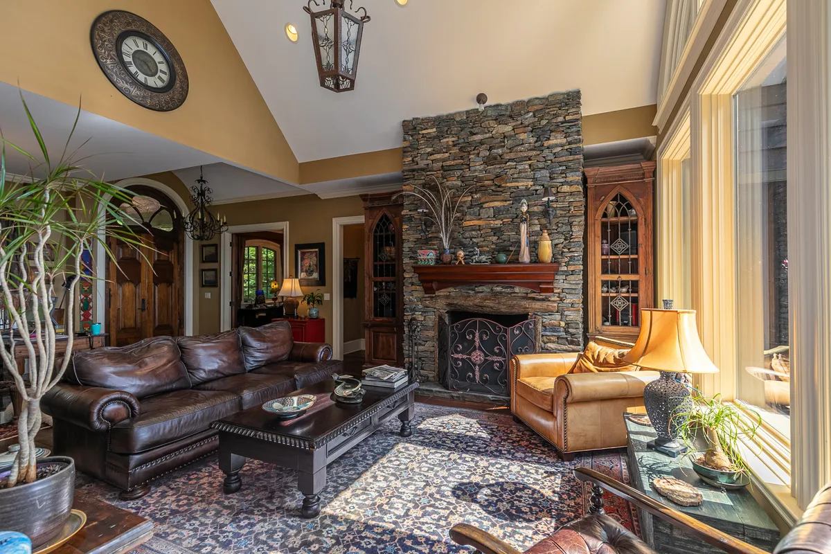 A spacious great room featuring a stacked-stone wood-burning fireplace, large arched windows framing panoramic mountain vistas, luxurious leather sofas, and decorative plants. The room is filled with natural light and exudes comfort and grandeur.